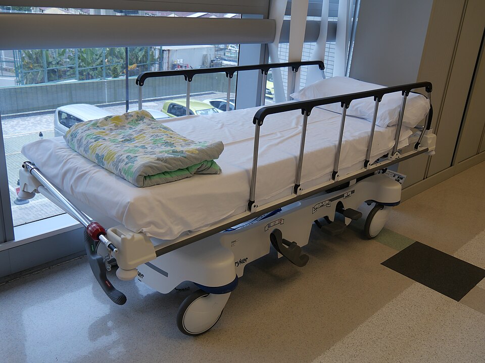An unoccupied wheeled hospital bed with raised side rails stands beside a window.
